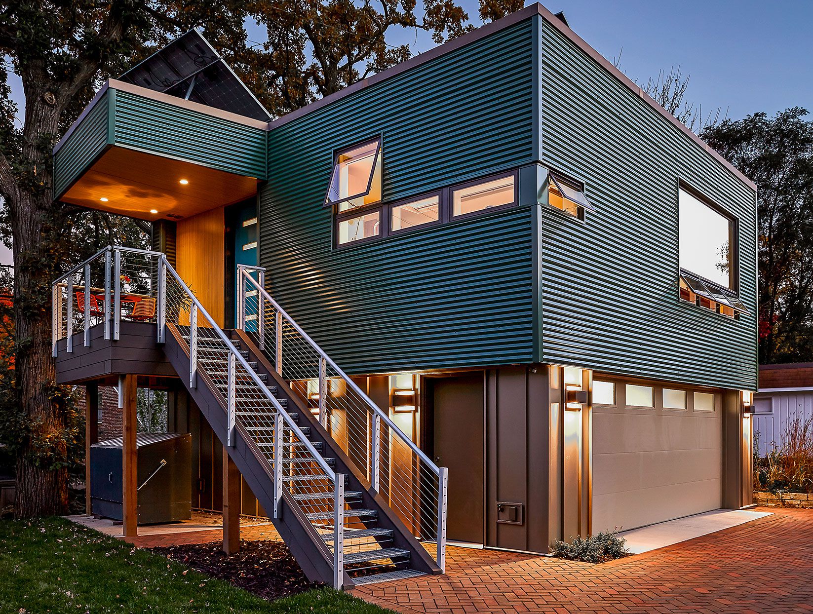 GargaeConv_ADU_AboveGarage Modern two-story house with blue corrugated metal siding, elevated entry via stairs, large garage door, and outdoor lighting, set against a backdrop of trees and evening sky.
